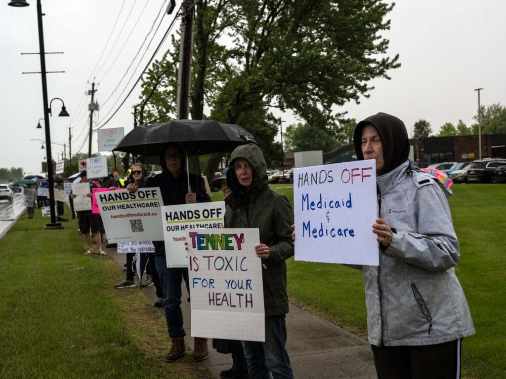 Protesters holding signs against healthcare cuts in rain.