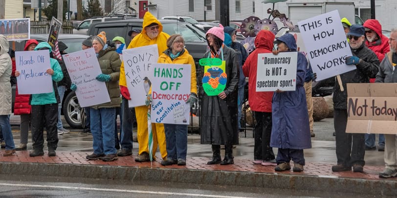 People standing on a sidewalk in raincoats with protest signs. Signs say things like "Hands Off Democracy," "Democracy is not a spectator sport," "This is How Democracy Works."