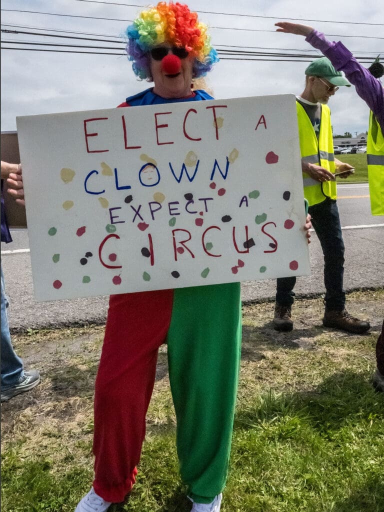 Person in clown outfit holding protest sign outdoors.