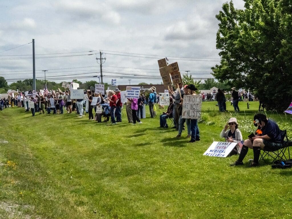 Crowd holds protest signs on grassy area.