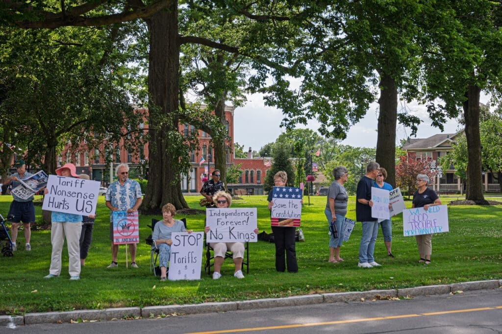 Protesters holding signs against tariffs on grassy area.