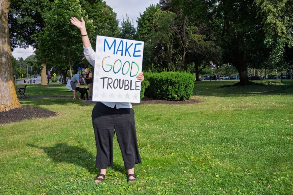 Person holds 'Make Good Trouble' sign in park.