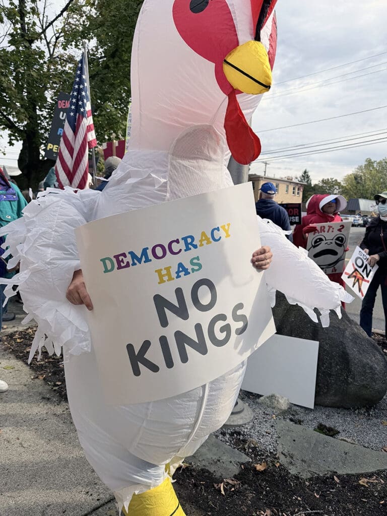 Person in chicken costume with 'Democracy Has No Kings' sign.