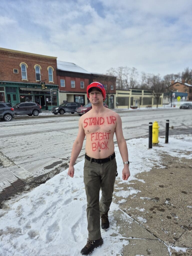 Man walks shirtless in snowy street with message.