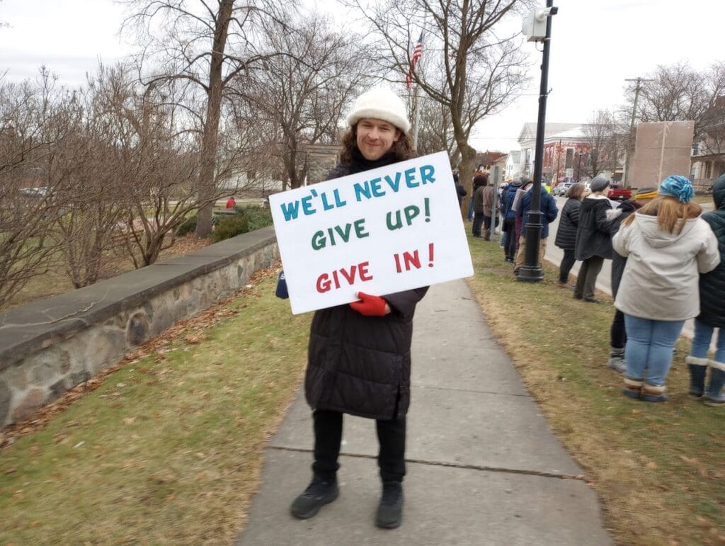 Person holding protest sign on sidewalk.