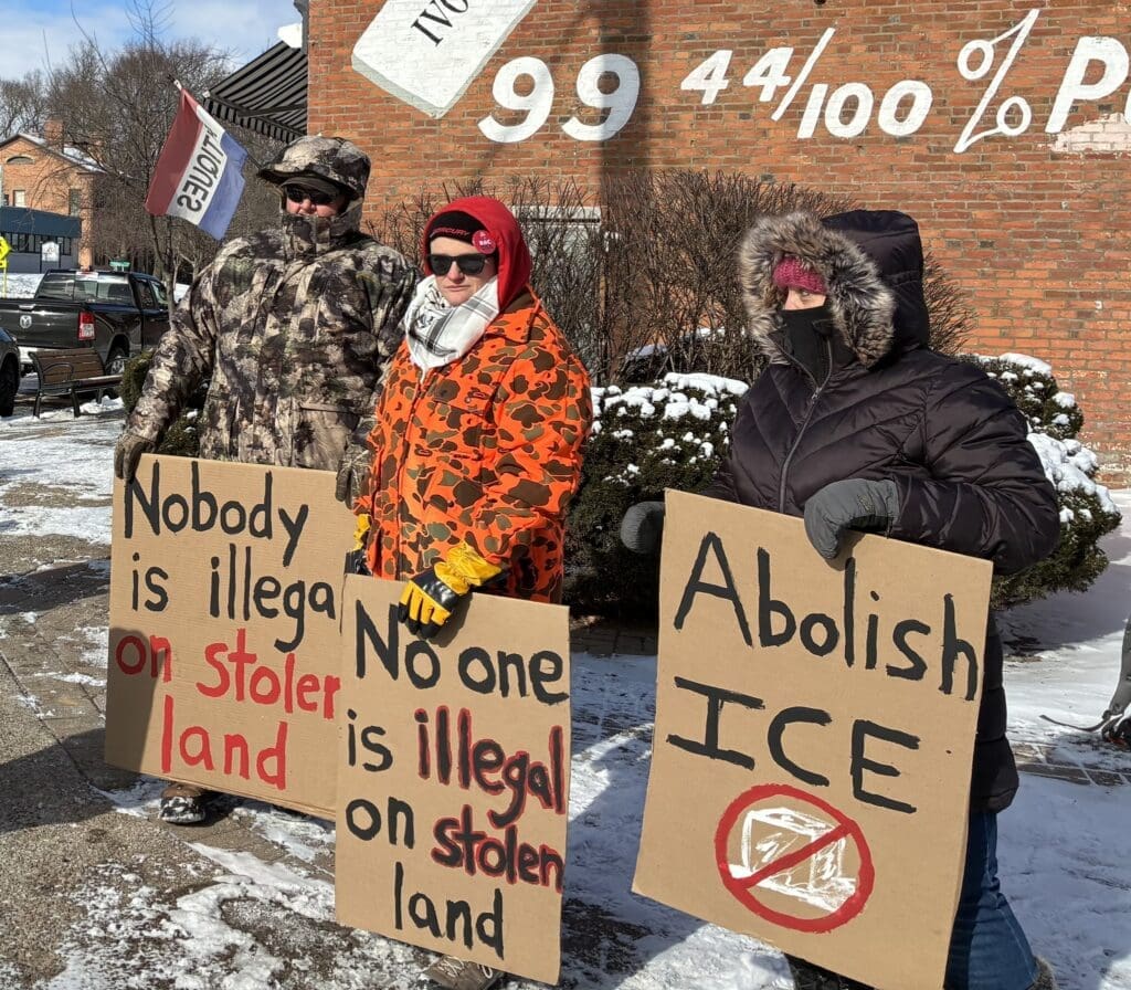 Protesters hold signs against ICE in snowy setting.