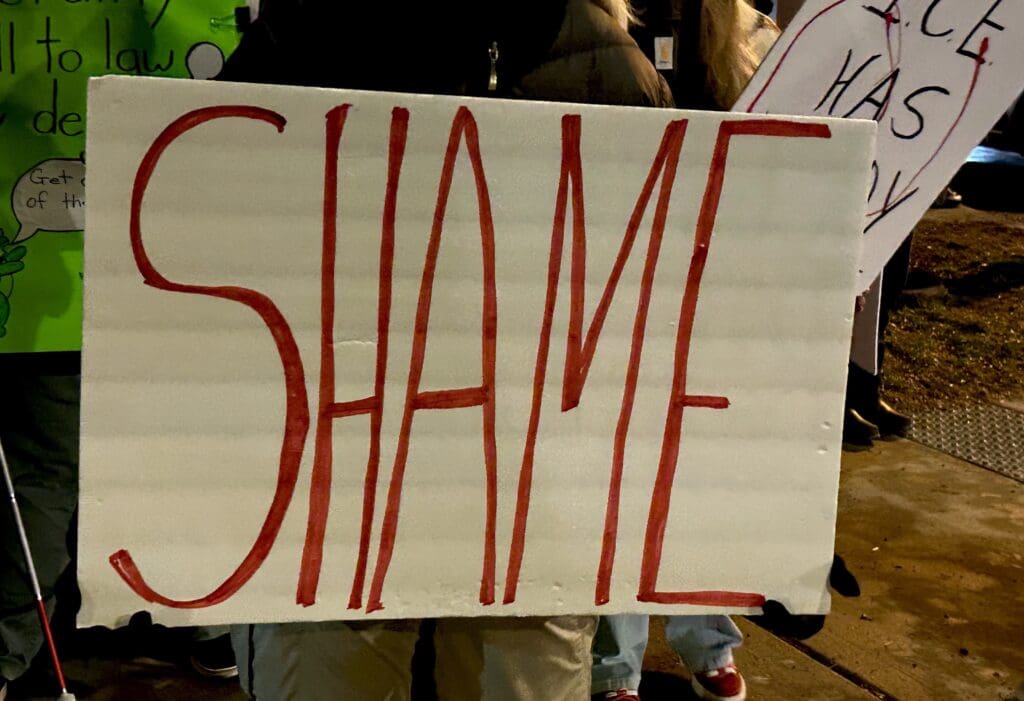 Protest sign with bold red 'SHAME' lettering.