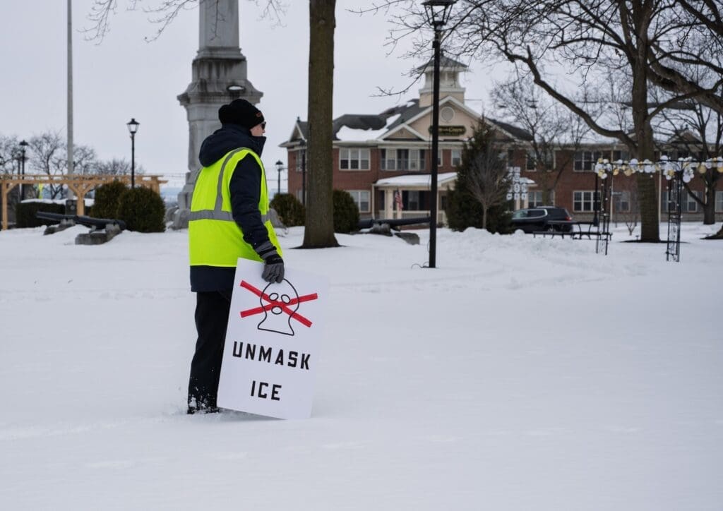 Person holding 'Unmask ICE' sign in snowy park.