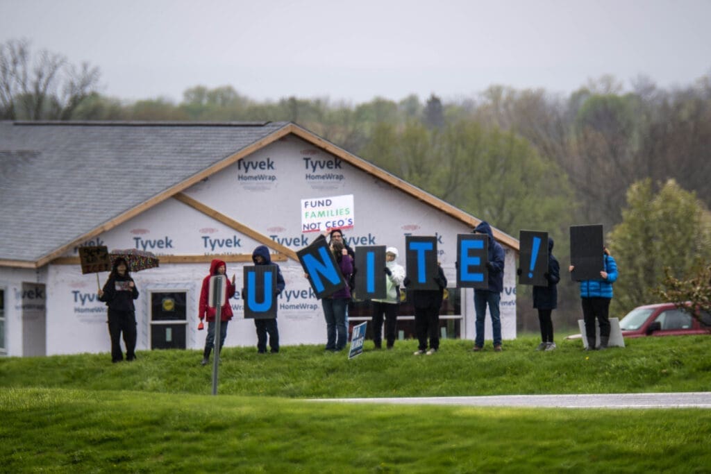 Protesters stand outside a house under construction holding large letters that spell "UNITE!" A nearby sign reads "Fund Families Not CEO's," calling for support for working families.