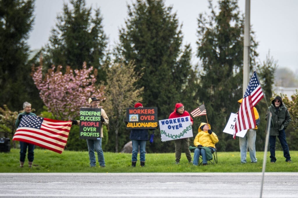 A small group of demonstrators gather with American flags and signs reading "Workers Over Billionaires" and "People Over Profits." The peaceful protest highlights calls for economic fairness and workers’ rights.