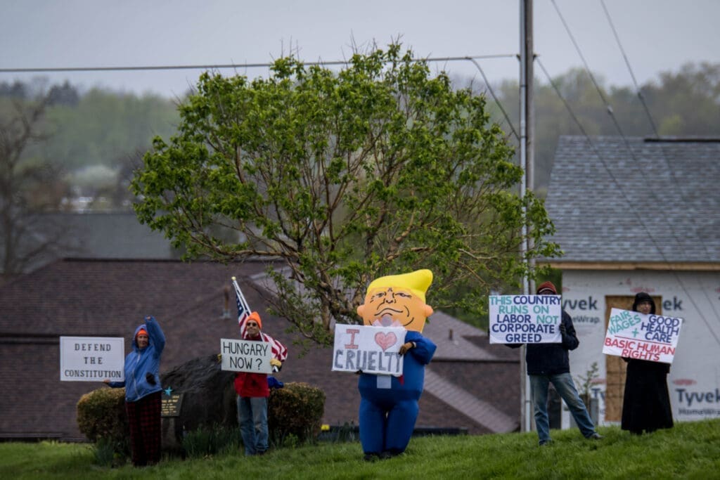 Protesters gather in a neighborhood holding signs supporting labor rights and the Constitution, alongside an oversized political caricature. The demonstration highlights concerns about corporate greed, healthcare, and basic human rights.