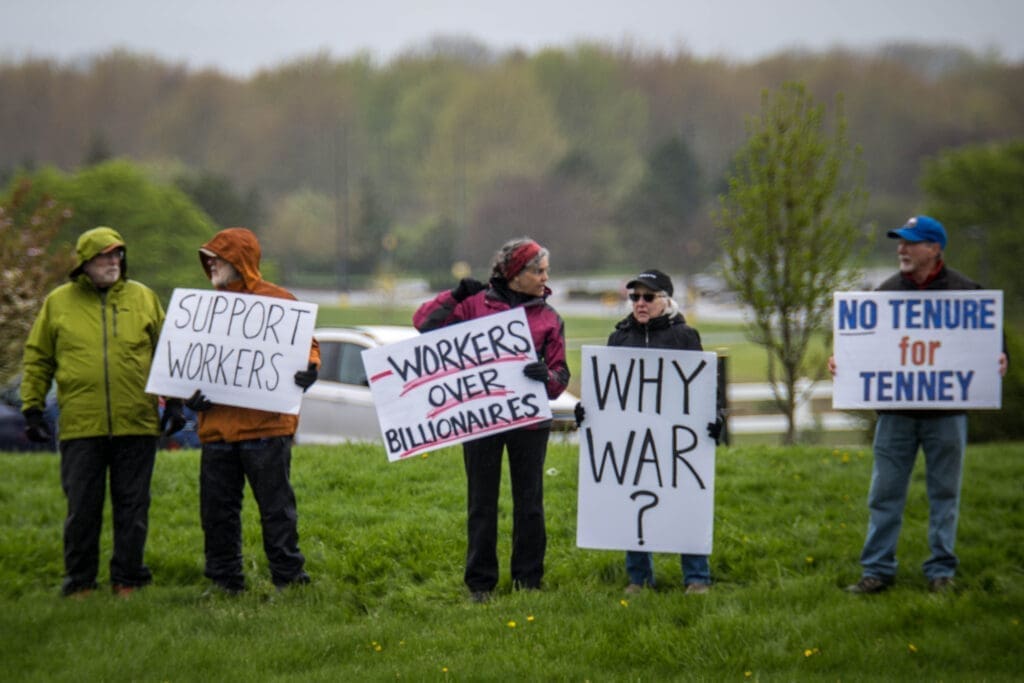 A small group of protesters stands outdoors holding signs advocating for workers’ rights and questioning political decisions. Their messages call for support of workers over billionaires and challenge local leadership.