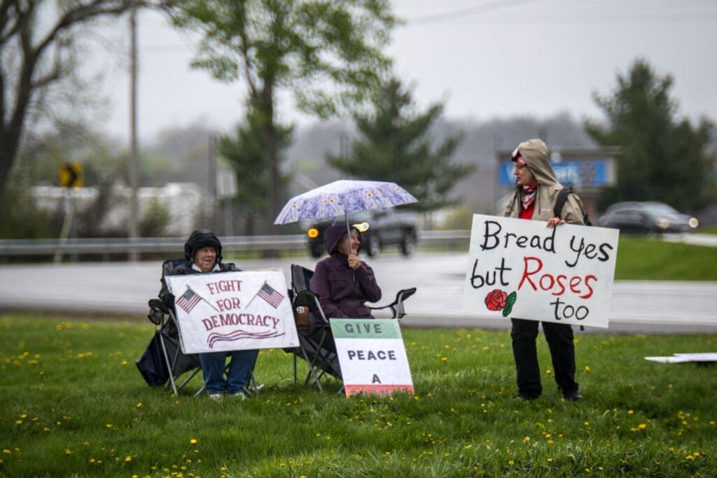 Protesters gather roadside in the rain, holding signs that call for democracy, peace, and dignity. Even under gray skies, their message stands bright and resolute.