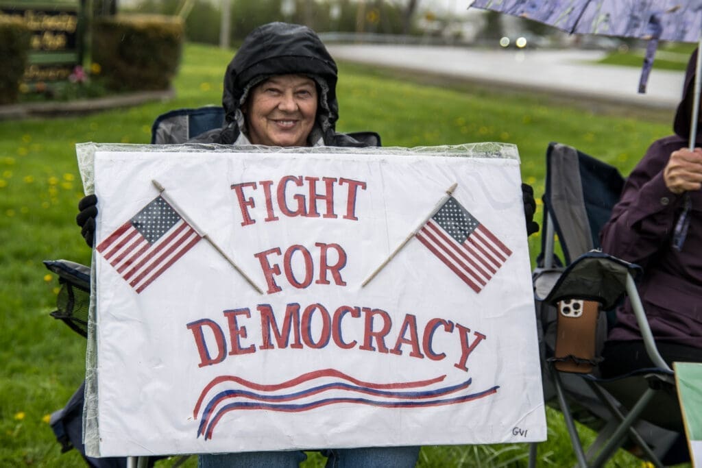 A smiling woman braves the rain while holding a "Fight for Democracy" sign adorned with American flags. Her determined spirit shines through at a peaceful outdoor rally.