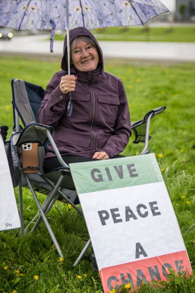 Smiling under an umbrella, a woman sits on the grass with a sign that reads "Give Peace a Chance." A quiet moment of hope and peaceful protest.