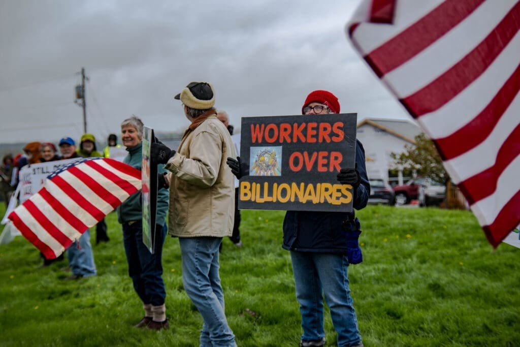 Protesters gather outdoors waving American flags, with one demonstrator holding a sign that reads "Workers Over Billionaires." The rally highlights calls for economic justice and workers’ rights.
