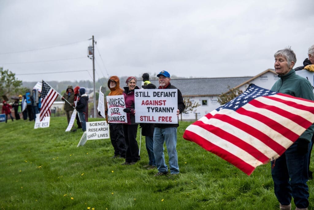 Protesters gather on a grassy field holding American flags and signs with messages about freedom, workers’ rights, and science.
