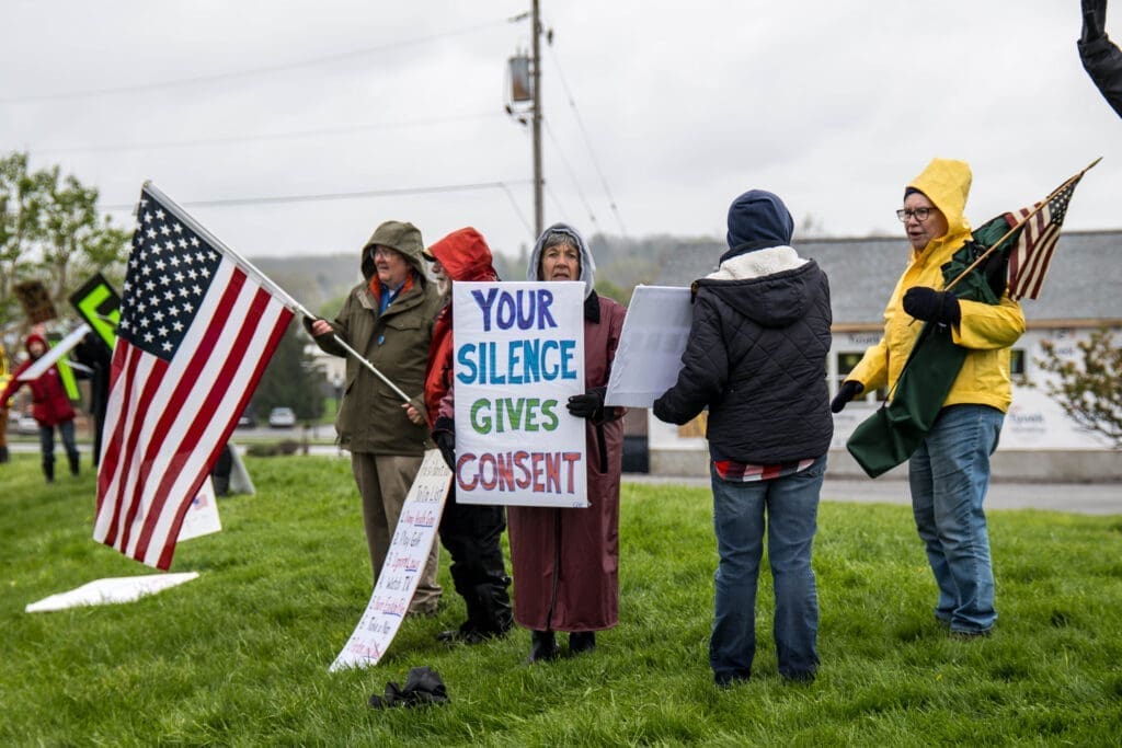 A small group of demonstrators stand in the rain holding American flags and protest signs, including one that reads "Your Silence Gives Consent." The gathering appears to take place along a roadside as they share their message with passing traffic.