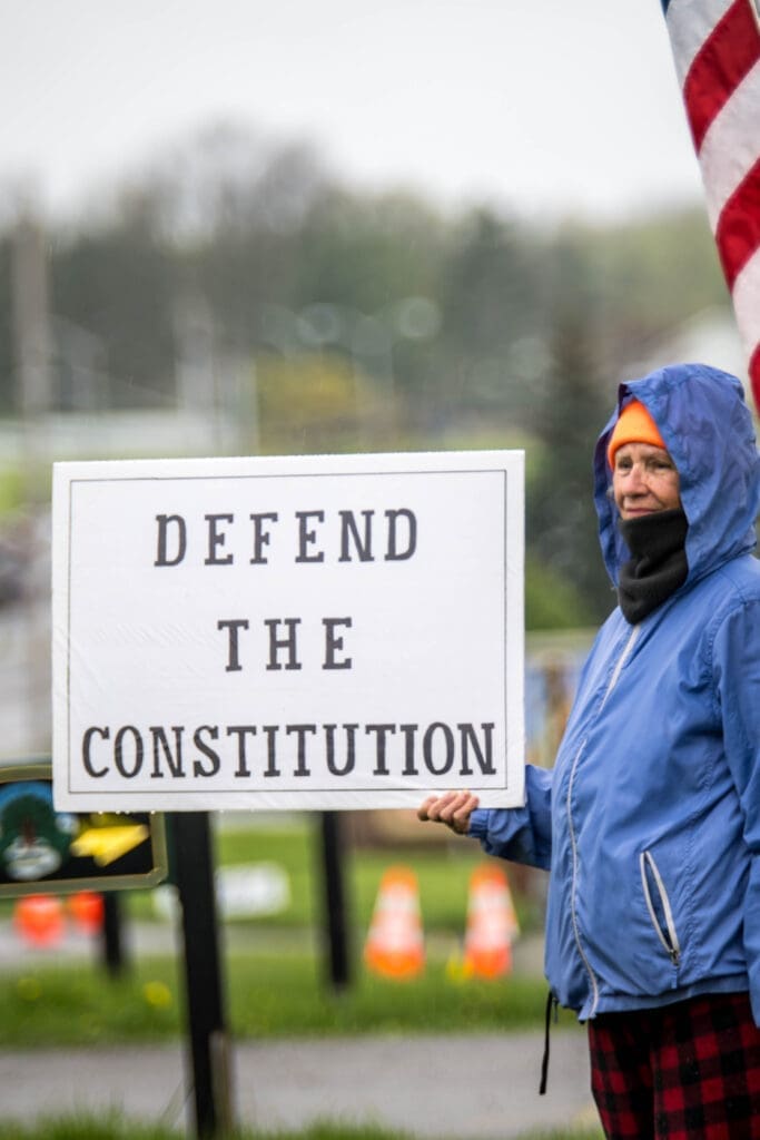 Protesters gather on a grassy field holding American flags and signs with messages about freedom, workers’ rights, and science. The small crowd stands united despite the gray, rainy weather.
