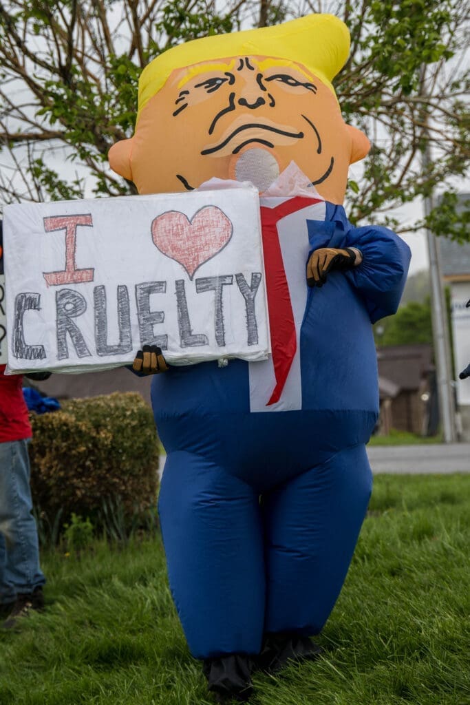 An inflatable caricature of a politician stands on a lawn holding a sign that reads "I ♥ Cruelty." The eye-catching display appears to be part of a protest or public demonstration.