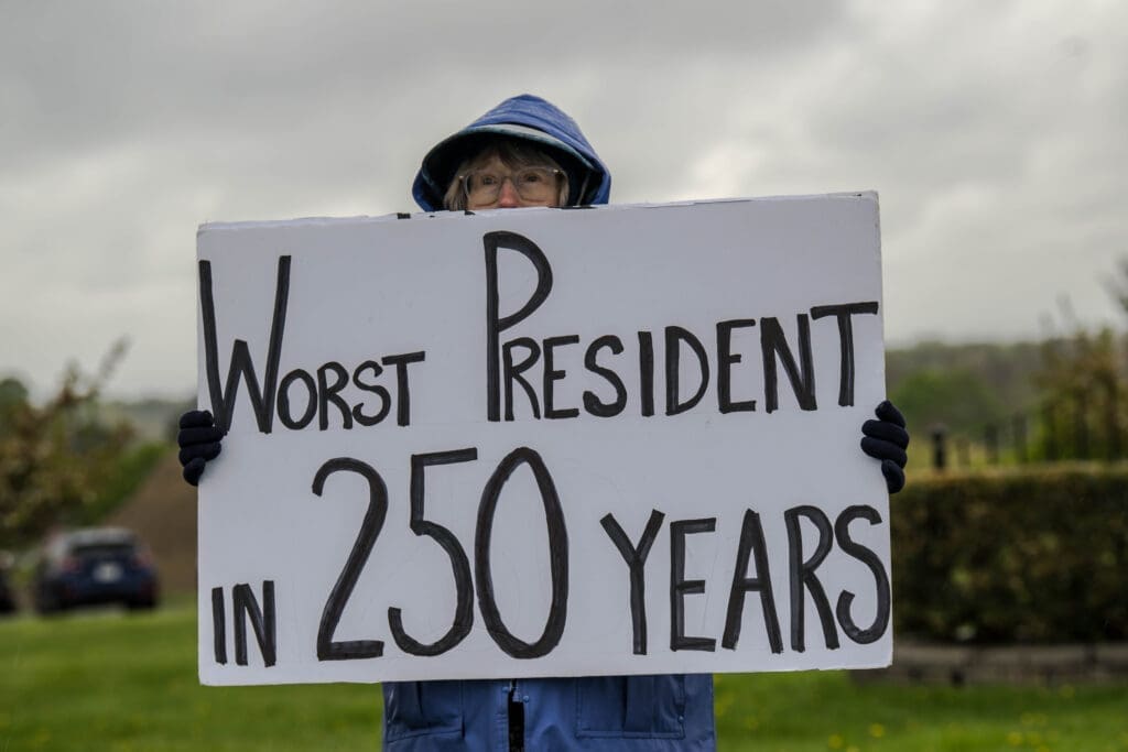 A bundled-up protester holds a bold sign reading "Worst President in 250 Years" on a cloudy day. The striking message captures a moment of political dissent and frustration.