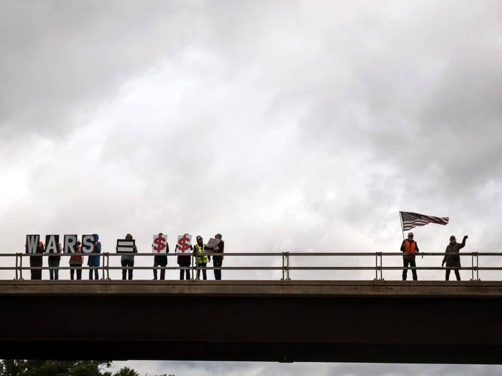 Protesters on bridge holding 'Wars Equal Money' signs and American flag