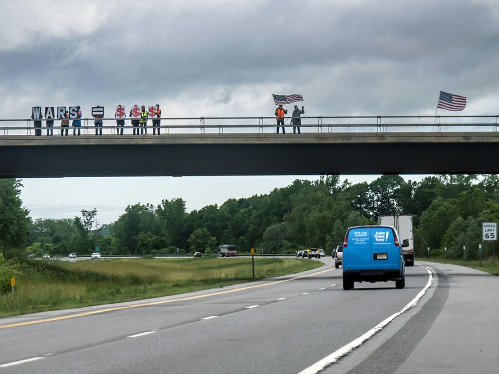 Protesters on highway overpass holding Wars Equal Money signs