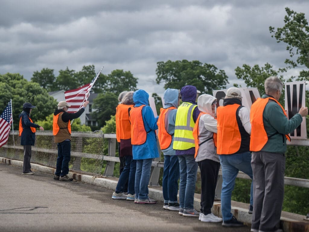 Group in orange vests holding signs and American flags on bridge